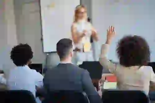 Photo of classroom, with students looking at teacher who is presenting in front of a whiteboard